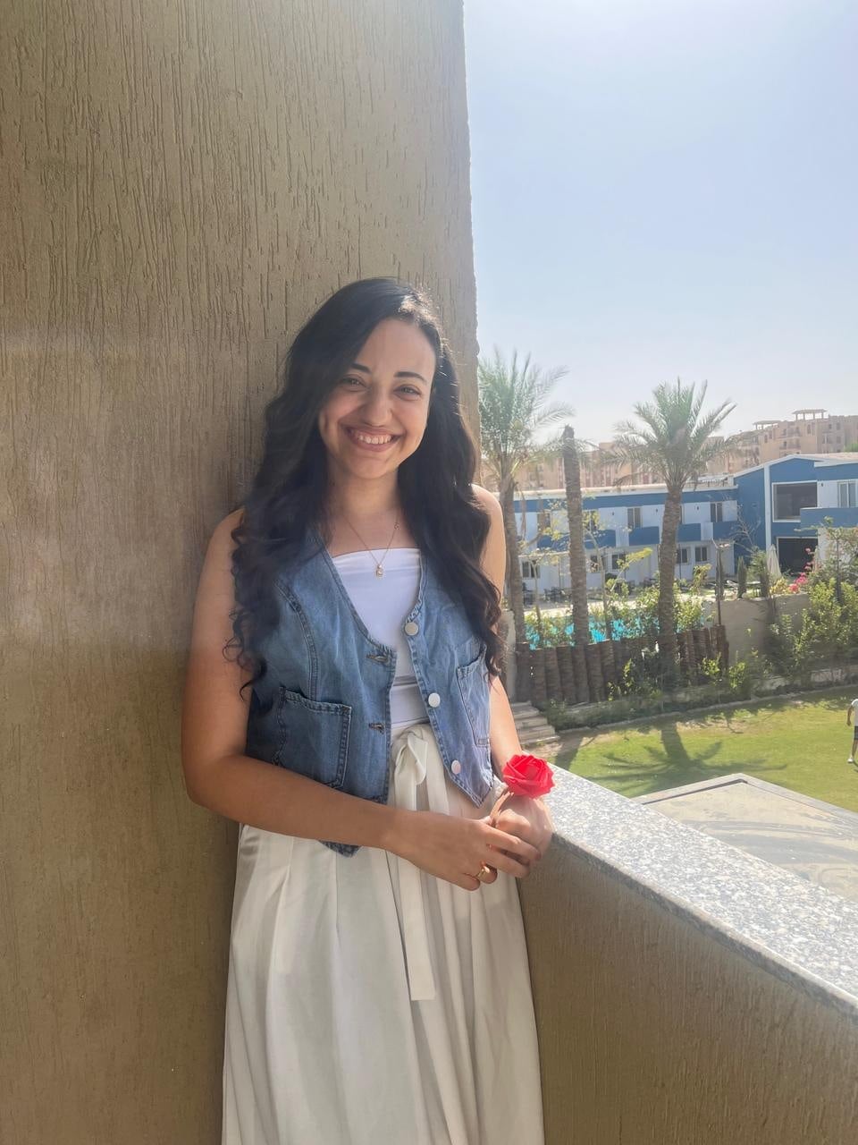 Nada, smiling, standing on a balcony in a white dress and denim vest